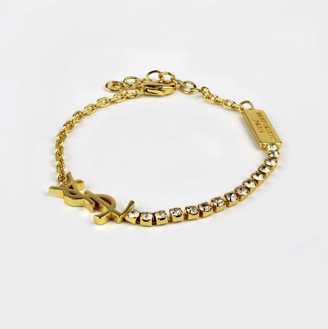 A gold-tone chain bracelet featuring a YSL logo charm, a row of clear rhinestones, and a small rectangular branded tag, photographed on a clean white background.