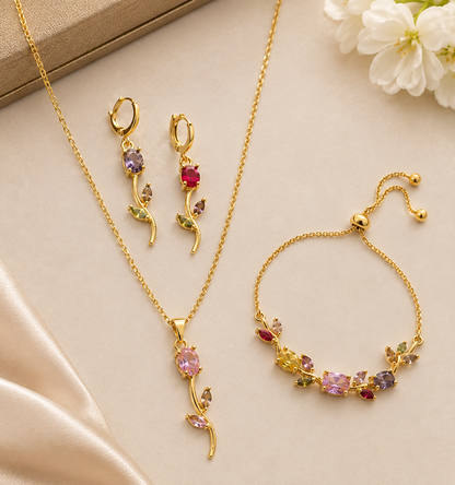 Gold jewelry set with a necklace, matching drop earrings, and a bracelet, decorated with colorful gemstones in pink, purple, red, and green, arranged on a beige satin background with white flowers.