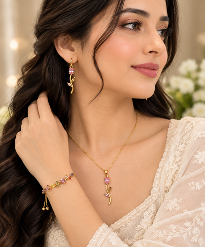 A woman wearing a gold jewelry set featuring a delicate necklace, matching drop earrings, and a bracelet with pink gemstone accents and gold leaf details.