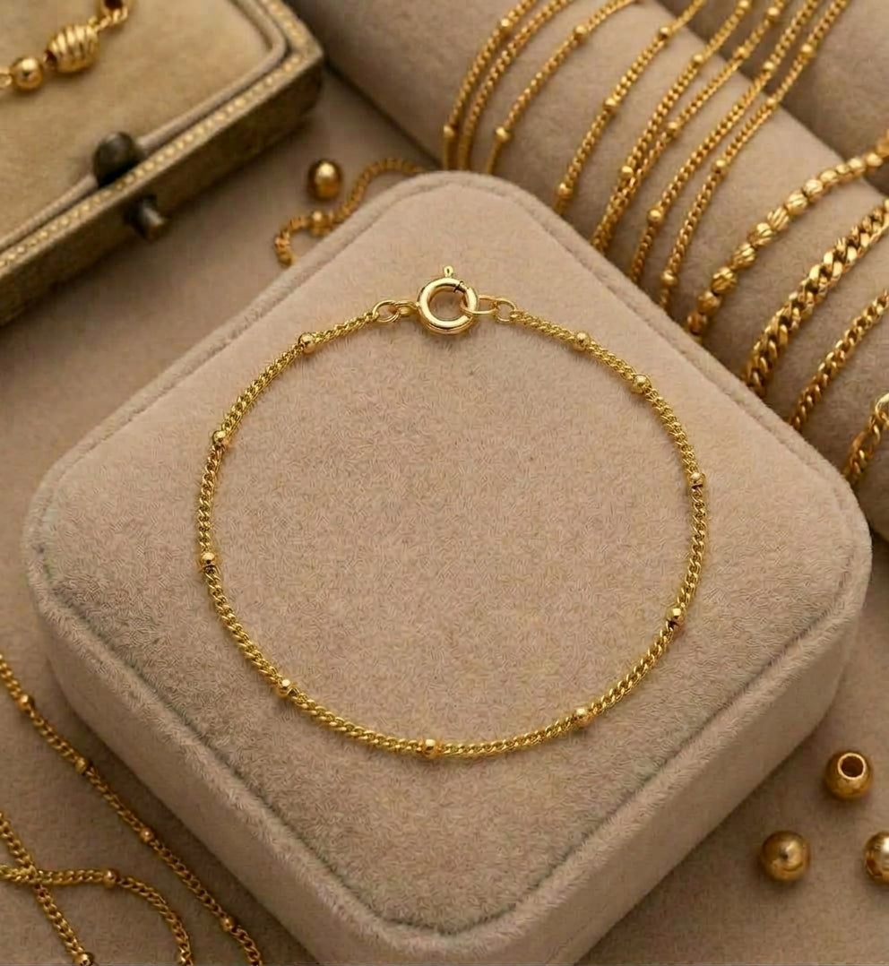 A delicate gold chain anklet with small bead accents and a circular clasp, displayed on a beige jewelry box. Additional gold necklaces and bracelets are arranged on display rolls in the background.