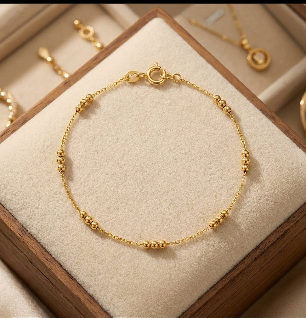 A delicate gold chain bracelet with small bead accents, displayed on a beige jewelry box cushion with other gold jewelry visible in the background.