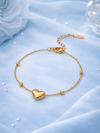 A delicate gold chain bracelet with a polished heart-shaped charm and small bead accents, displayed on a soft light blue fabric background.