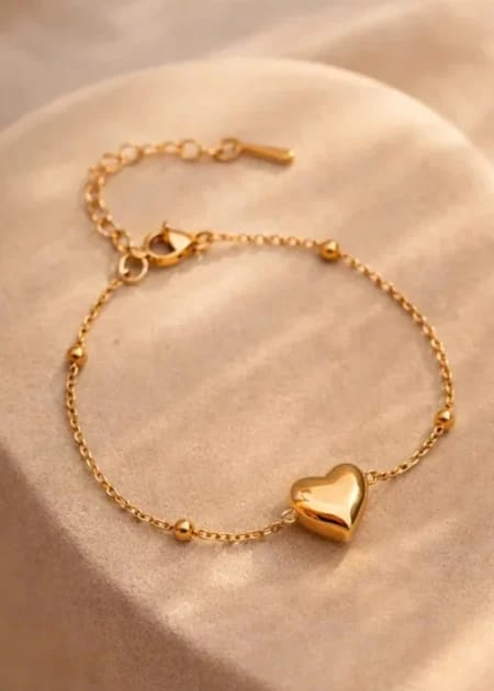 A delicate gold chain bracelet with a polished heart-shaped charm and small bead accents, displayed on a soft beige fabric surface.