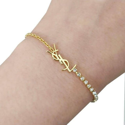 A gold bracelet with a YSL logo charm and a row of clear stones, shown worn on a person's wrist against a white background.