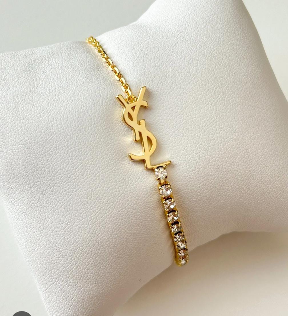 A gold-tone YSL bracelet with a rhinestone-embellished bar, displayed on a white textured jewelry stand.
