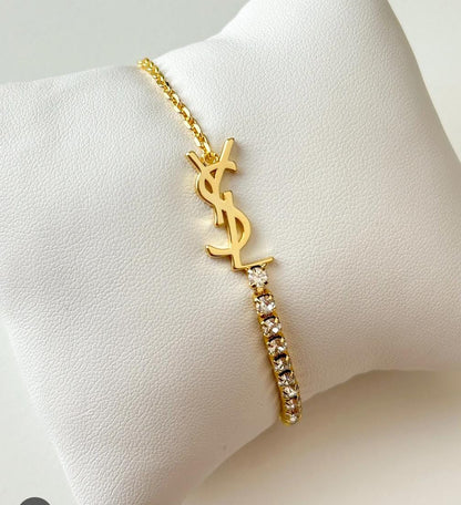 A gold-tone YSL bracelet with a rhinestone-embellished bar, displayed on a white textured jewelry stand.