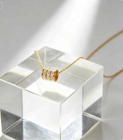 A gold necklace with a cylindrical pendant set with clear stones, displayed on a transparent acrylic block against a bright white background with soft shadows.