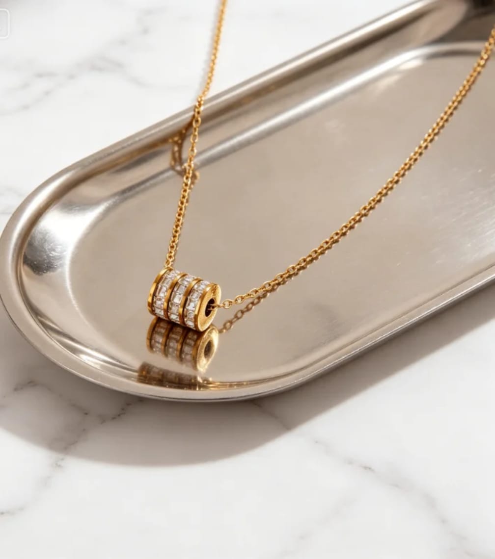 A gold chain necklace with a cylindrical pendant featuring three rows of clear stones, displayed on a polished silver tray over a white marble surface.