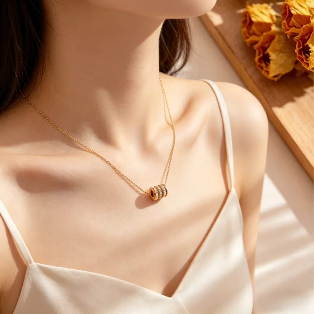 A close-up lifestyle shot of a woman wearing a delicate gold chain necklace with a cylindrical pendant featuring three horizontal rows of small clear stones. She is wearing a cream-colored satin camisole, with warm sunlight casting soft shadows on her skin and a wooden tray with dried yellow flowers in the background.