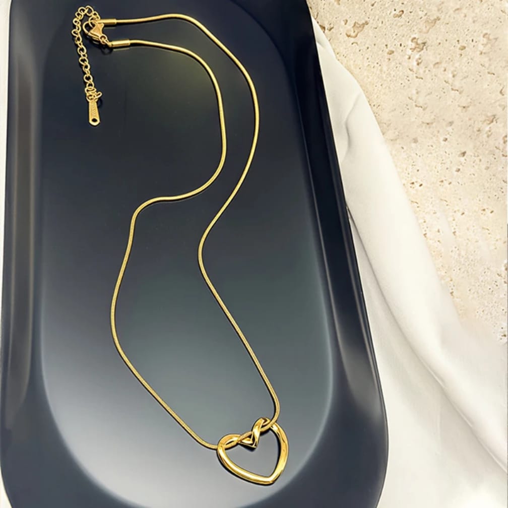 A gold-toned chain necklace with a double-heart pendant, displayed on a dark gray tray next to a white fabric and textured beige surface.