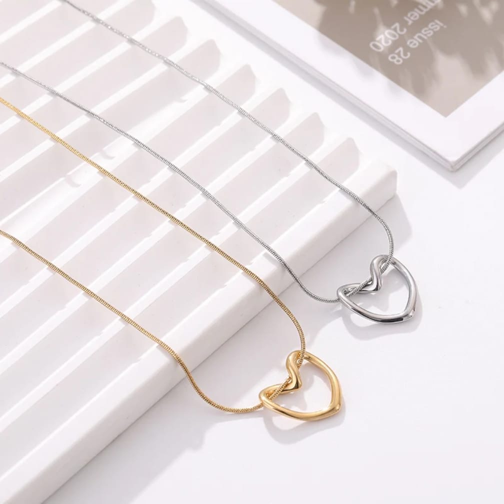 Two interlocking heart pendant necklaces on delicate chains, one in polished gold and one in polished silver, displayed on a white slatted tray with a magazine in the background.