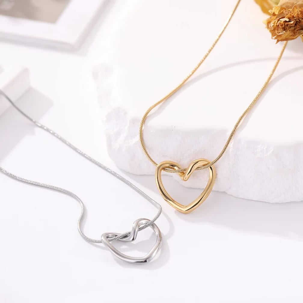 Two interlocking heart-shaped necklaces displayed on a clean white surface, one in polished gold and one in polished silver, with a white stone slab and dried flower as styling props.
