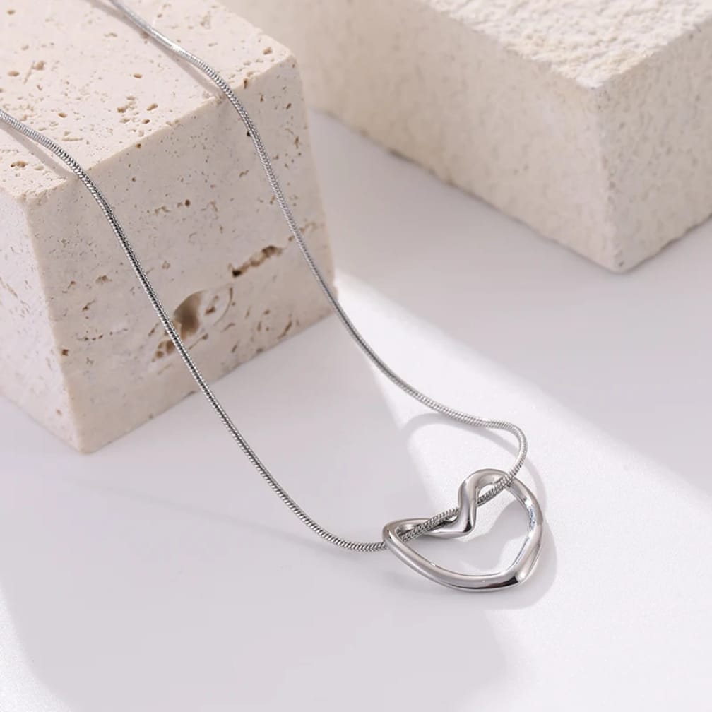 A polished silver necklace with an interlocking double-heart pendant, draped elegantly over a textured beige stone block on a clean white surface.