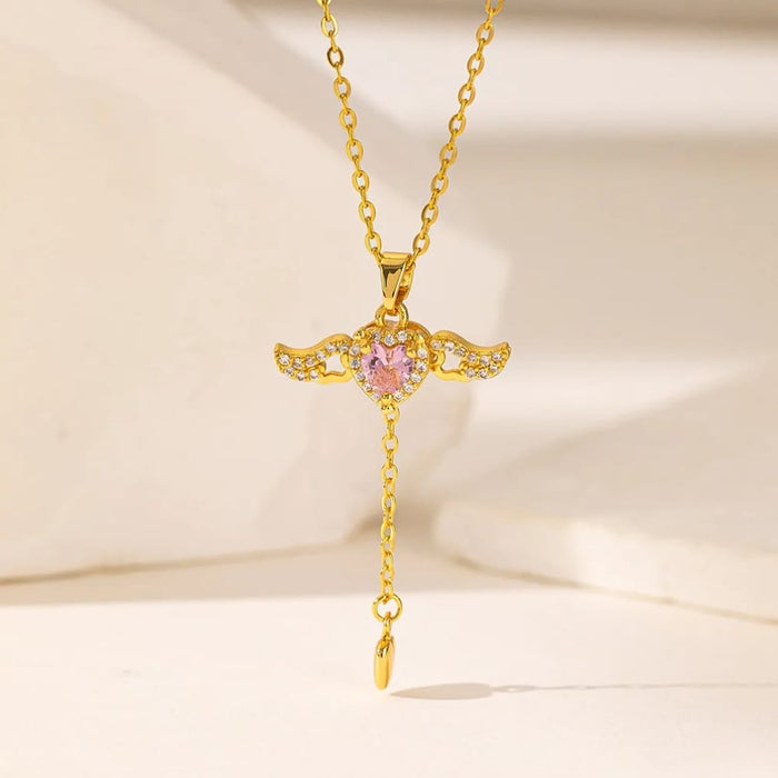 Gold angel wings pendant necklace with a pink gemstone and small clear crystals, shown on a delicate chain against a soft cream background.