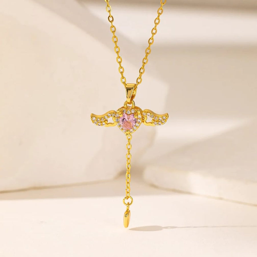 Gold angel wings pendant necklace with a pink gemstone and small clear crystals, shown on a delicate chain against a soft cream background.