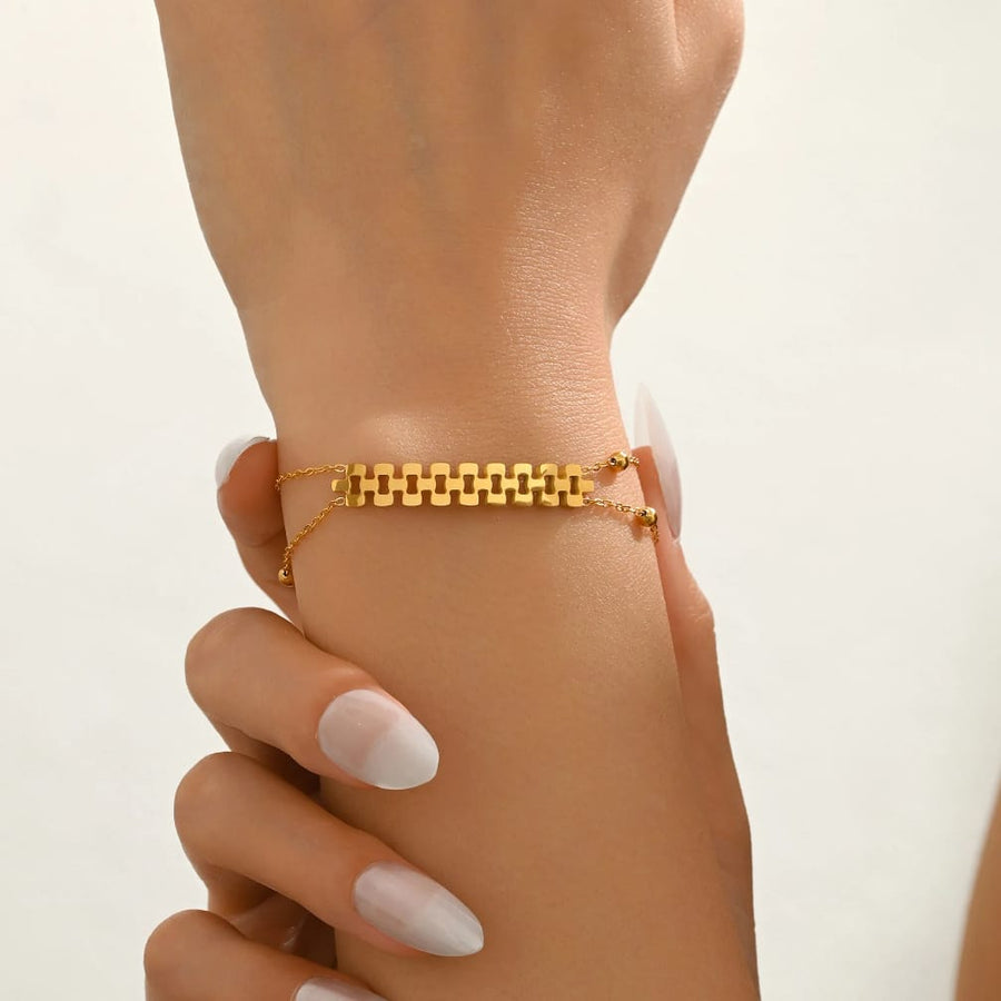 A delicate gold chain bracelet with a central geometric link pattern, shown worn on a person's wrist with manicured nails visible.