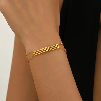A close-up of a delicate gold double-strand bracelet worn on a person's wrist. The bracelet features a central geometric link pattern on one side and a fine chain on the other, set against a background of skin tones and a black garment.