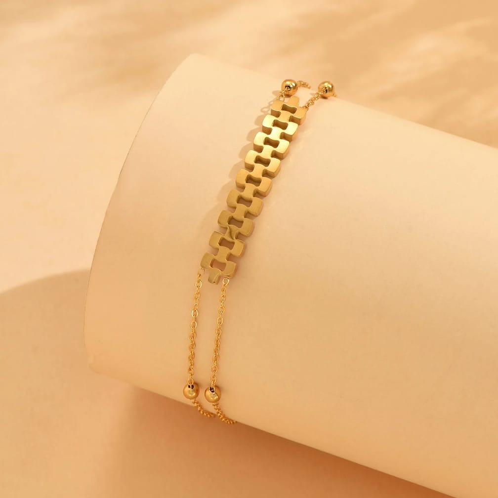 A gold-toned double-strand bracelet displayed on a cream-colored cylindrical jewelry stand against a warm beige background.