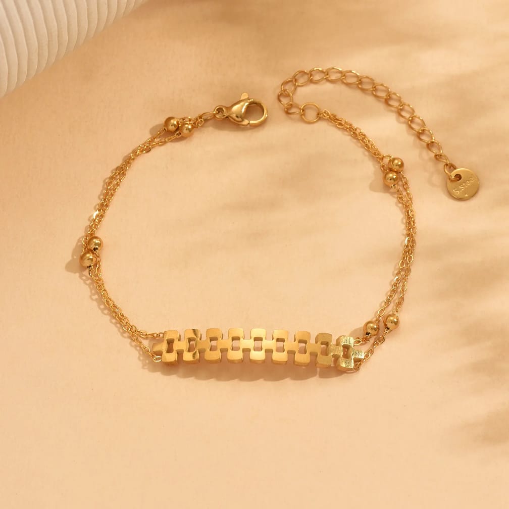 A delicate gold chain bracelet with a central horizontal bar made of repeating rectangular links, accented by small gold beads, shown on a warm beige fabric background.
