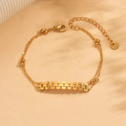 A delicate gold chain bracelet with a central horizontal bar made of repeating rectangular links, accented by small gold beads, shown on a warm beige fabric background.