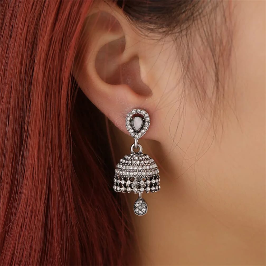 A close-up of a person's ear wearing a silver-toned jhumka earring adorned with clear stones and dangling beadwork, against red hair and skin.