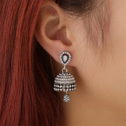 A close-up of a person's ear wearing a silver-toned jhumka earring adorned with clear stones and dangling beadwork, against red hair and skin.