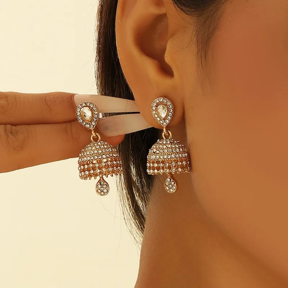 A close-up of a person wearing gold-plated jhumka earrings adorned with clear stones, with a hand holding the earring on the other side for comparison against a warm beige background.