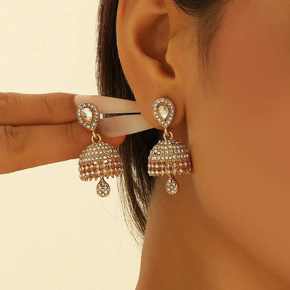 A close-up of a person wearing gold-plated jhumka earrings adorned with clear stones, with a hand holding the earring on the other side for comparison against a warm beige background.