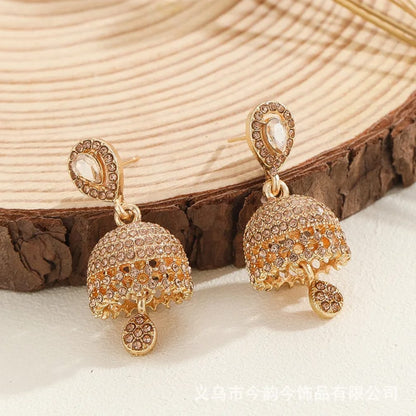 A pair of gold-toned Indian jhumka earrings adorned with clear rhinestones and small amber-colored stones, displayed on a rustic wood slice background.