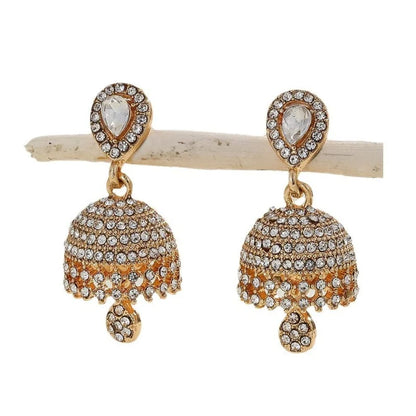 A pair of gold-plated jhumka earrings adorned with clear crystals, displayed on a small natural wood branch against a plain white background.