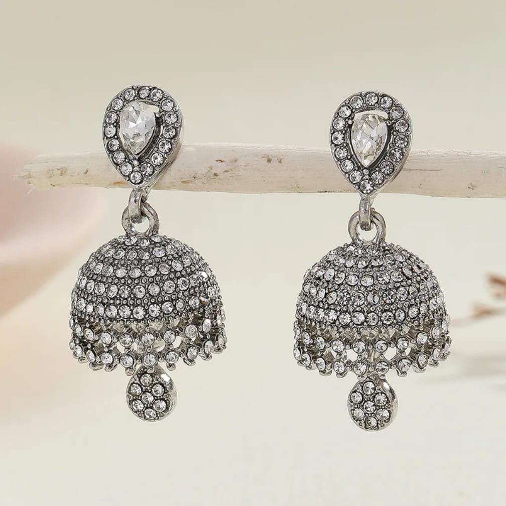 A pair of silver-toned jhumka earrings adorned with clear pavé rhinestones, featuring a teardrop-shaped clear stone at the top and a bell-shaped dangling cluster with a small round stone at the bottom, displayed on a light wooden branch against a soft cream background.