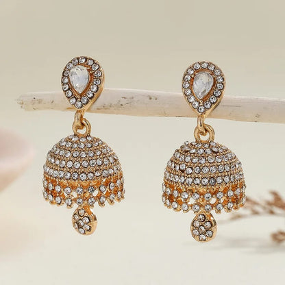 A pair of gold-plated Indian jhumka earrings adorned with clear crystals, displayed on a light wooden branch against a soft beige background.
