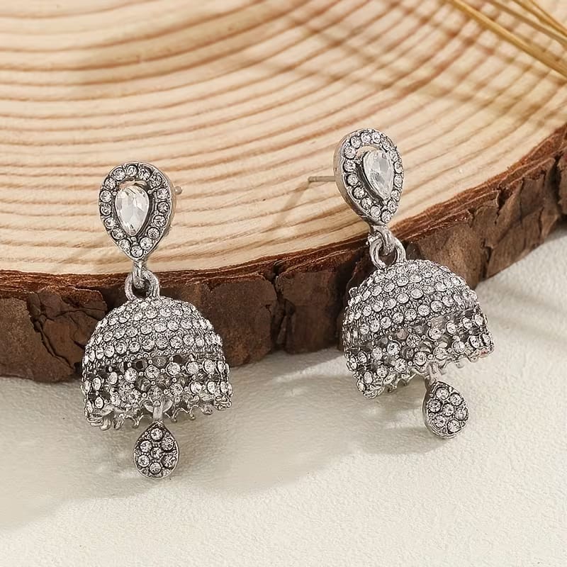 A pair of silver-toned jhumka earrings adorned with clear rhinestones, displayed on a rustic wood slice over a light beige surface.
