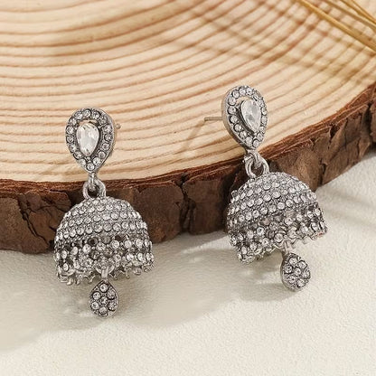 A pair of silver-toned jhumka earrings adorned with clear rhinestones, displayed on a rustic wood slice over a light beige surface.