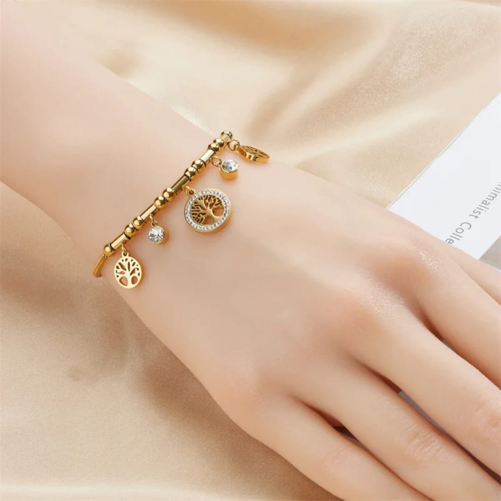 A gold-toned charm bracelet with a tree of life pendant, a sun face charm, and a small clear stone charm, shown worn on a hand against a soft beige satin background.