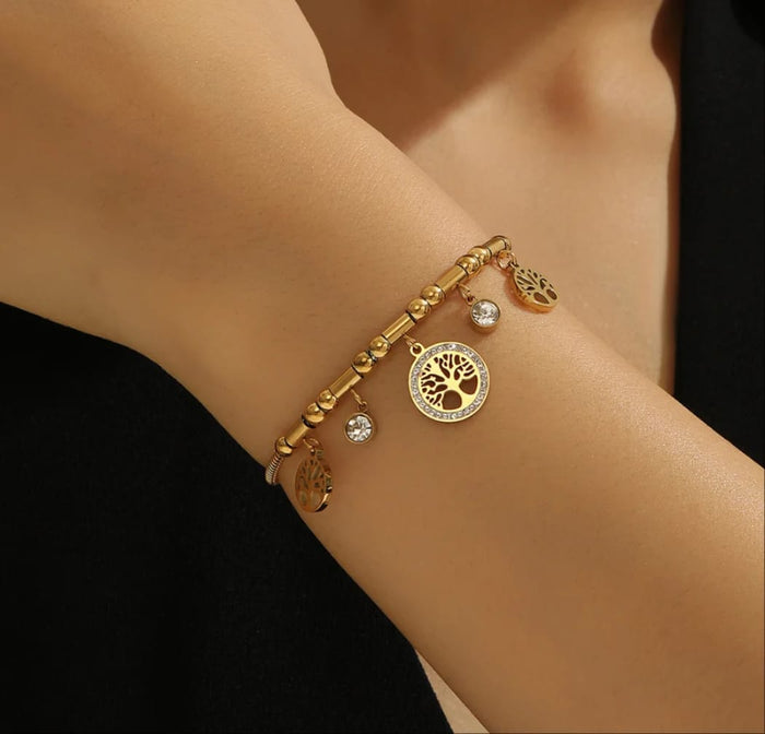 A gold-toned beaded bracelet with tree-of-life and leaf charms, worn on a person's wrist against a dark background.