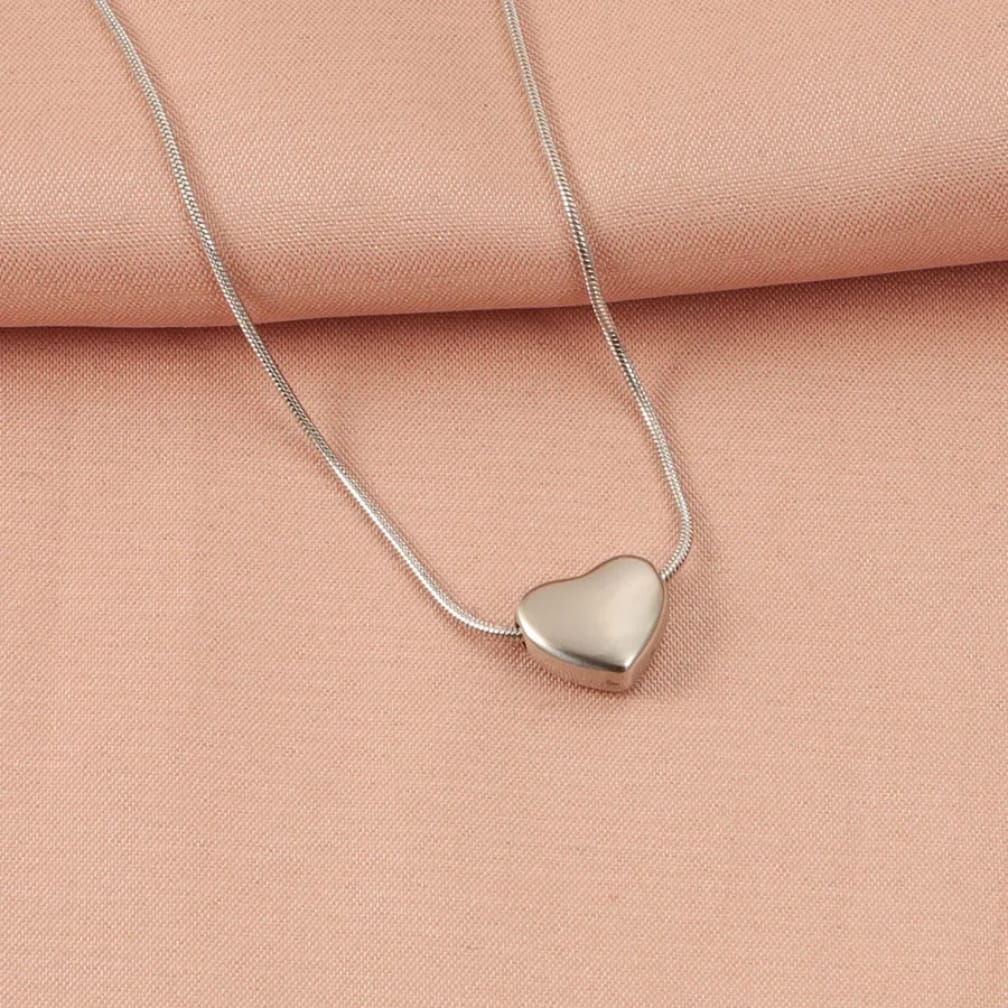 A silver snake chain necklace with a polished, heart-shaped pendant, displayed on a soft peach-pink textured fabric background.