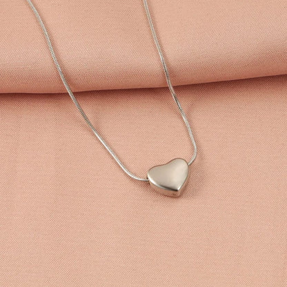 A silver snake chain necklace with a polished, heart-shaped pendant, displayed on a soft peach-pink textured fabric background.