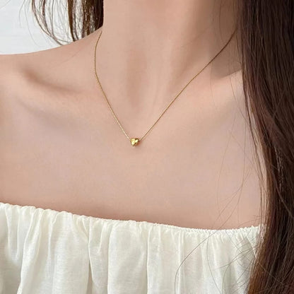 A delicate gold chain necklace with a small heart-shaped pendant, worn by a model in a close-up shot. The model is wearing a white off-the-shoulder top.