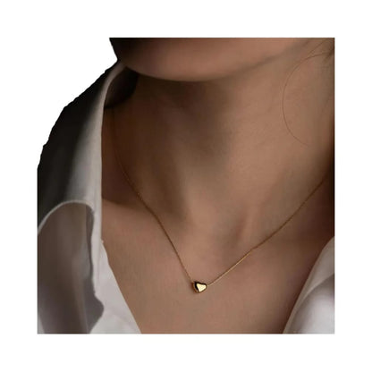 A close-up of a model's neck and collarbone area wearing a delicate gold chain necklace with a small, polished gold heart pendant, styled with a white collared shirt.