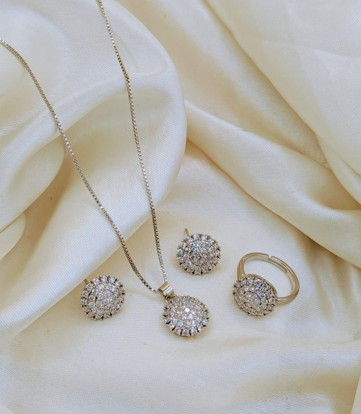 A gold jewelry set featuring a necklace, ring, and earrings with round pavé crystal pendants, arranged on draped cream-colored satin fabric.