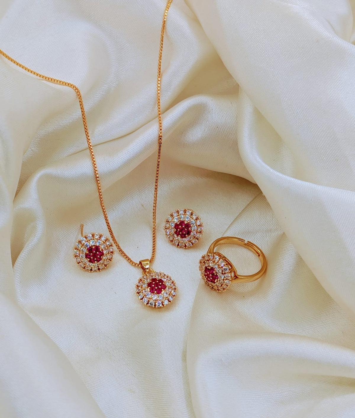 Gold jewelry set with a pendant necklace, matching earrings, and a ring, each featuring a central round red gemstone surrounded by small clear stones, displayed on draped cream satin fabric.