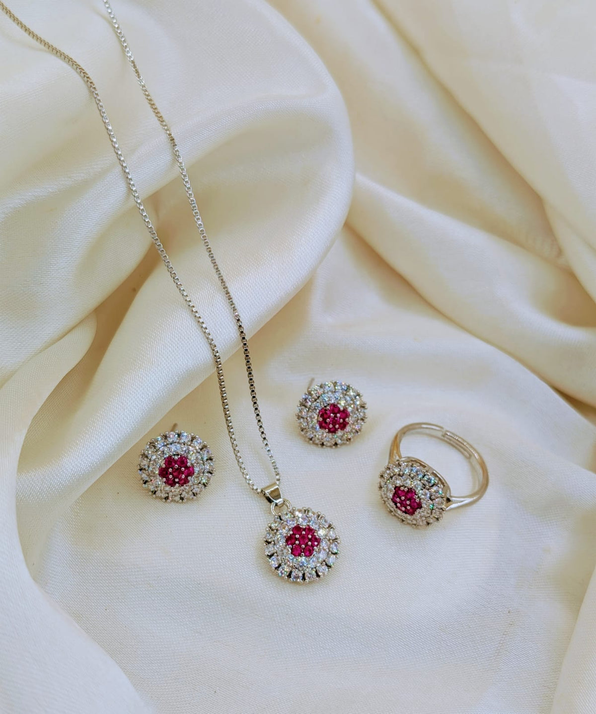 A gold jewelry set featuring a necklace, earrings, and ring with round pink gemstones surrounded by clear crystals, displayed on draped cream-colored satin fabric.