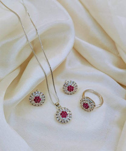 A gold jewelry set featuring a necklace, earrings, and ring with round pink gemstones surrounded by clear crystals, displayed on draped cream-colored satin fabric.