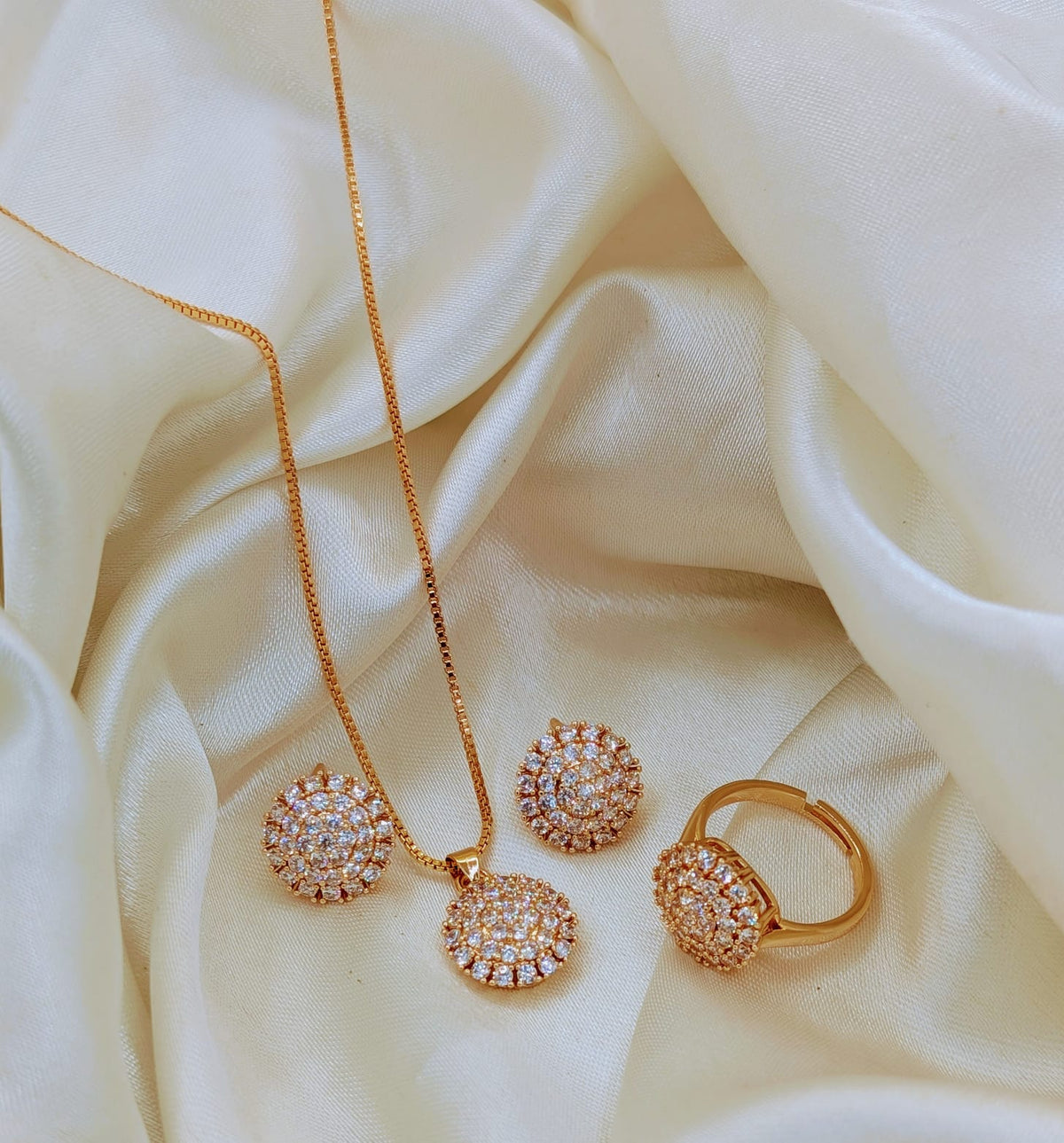 A gold jewelry set featuring a necklace, ring, and earrings, all adorned with sparkling clear pavé stones, arranged on a draped cream satin fabric background.
