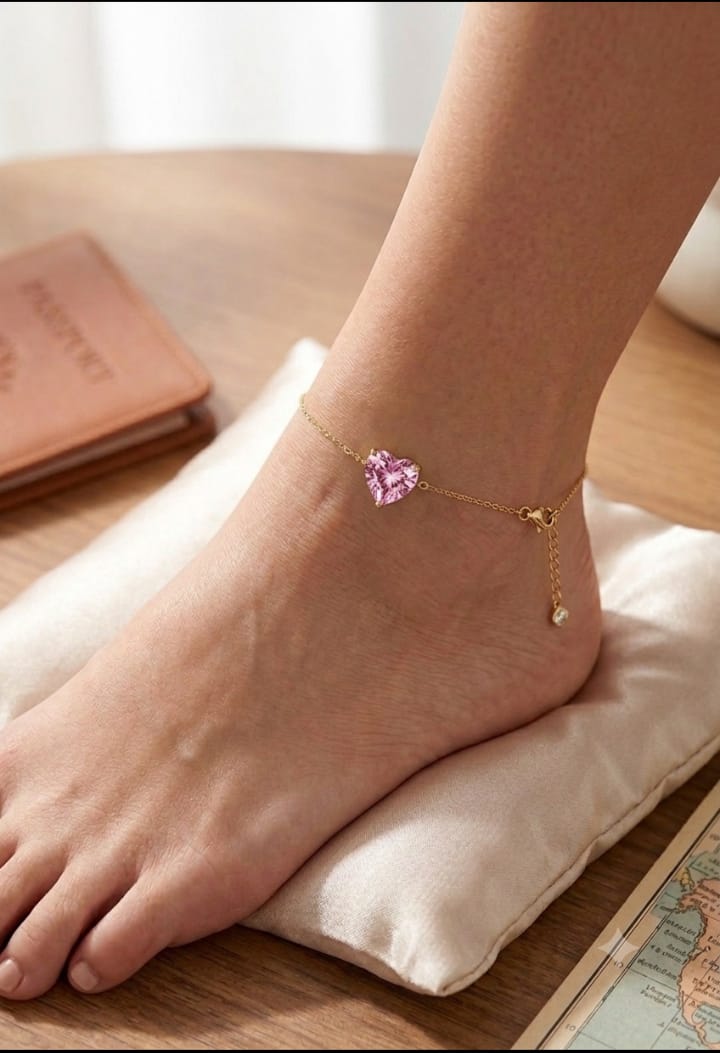 A delicate gold chain anklet with a small pink heart-shaped crystal charm is shown worn on a person's ankle, resting on a cream satin pillow over a wooden surface.