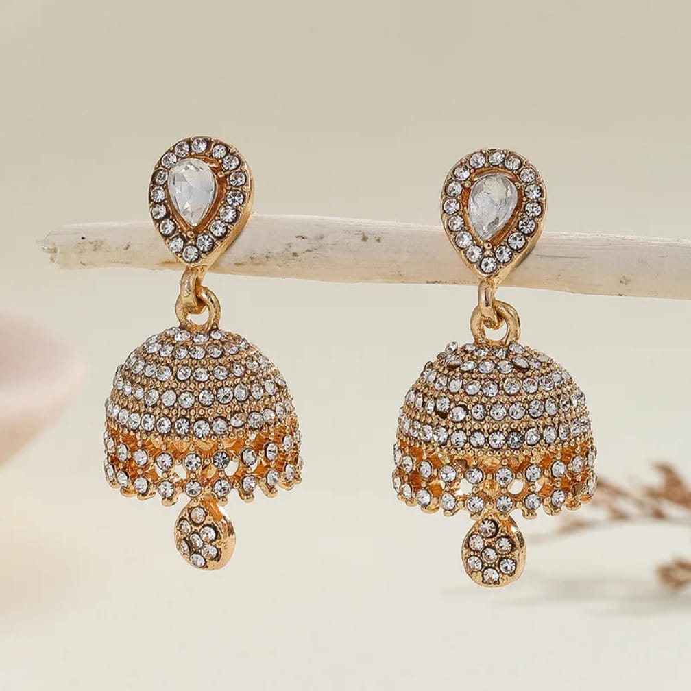 A pair of gold-plated Indian jhumka earrings adorned with clear crystals, displayed on a light wooden branch against a soft beige background.