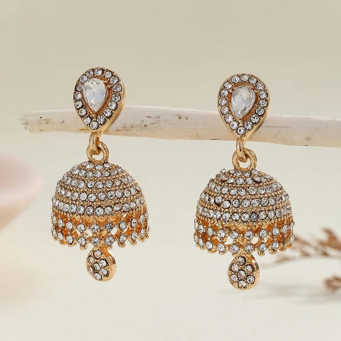 A pair of gold-plated Indian jhumka earrings adorned with clear crystals, displayed on a light wooden branch against a soft beige background.