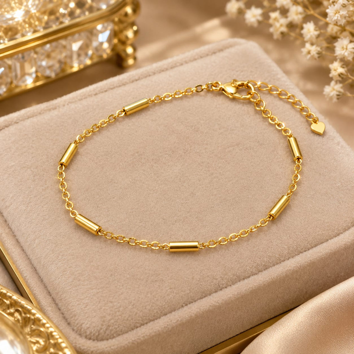 A gold chain bracelet with cylindrical links, displayed on a beige velvet jewelry box with decorative gold and white floral elements in the background.