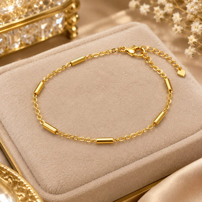 A gold chain bracelet with cylindrical links, displayed on a beige velvet jewelry box with decorative gold and white floral elements in the background.